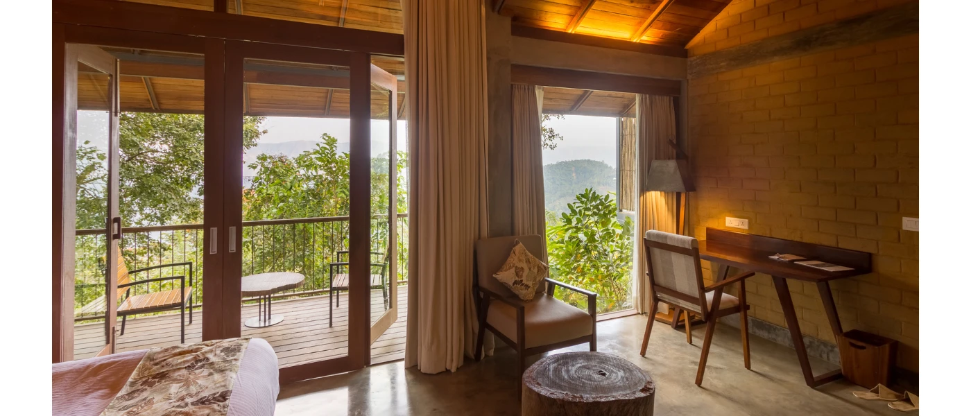 Hotel room with neutral colouring, desk space, a chair and coffee table and floor-to-ceiling patio doors opening onto a furnished terrace