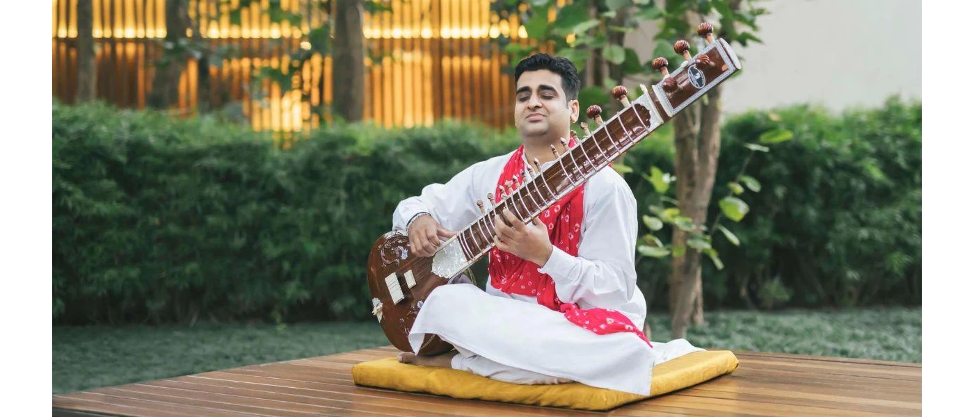 Man in white robe and red patterned scarf plays an instrument while sitting on a yellow cushion