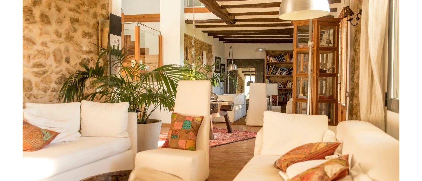 Cosy lounge area with white sofas, cobbled stone walls, wood-beamed ceilings, potted green plants and a bookcase filled with books