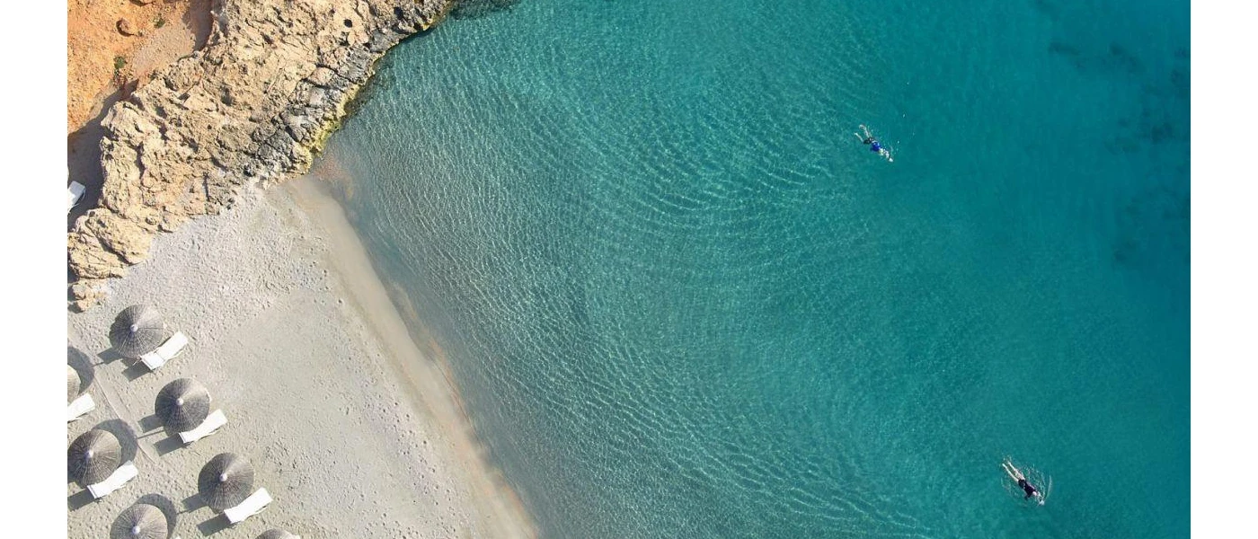 Sandy beach with white-cushioned loungers and thatched umbrellas, lapped by turquoise shallows and next to a craggy cliff