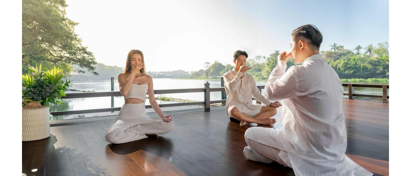 Three people in white sit cross-legged with their eyes closed holding their noses on a wooden deck next to a river