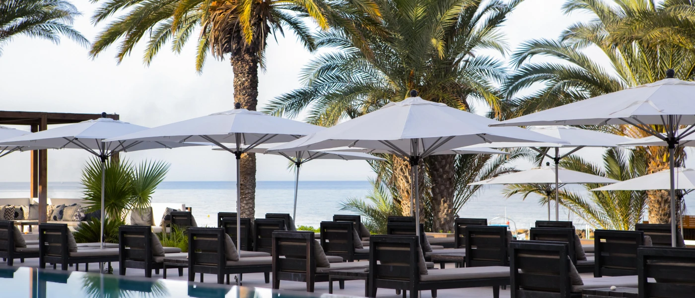 Cushioned wooden sun loungers in rows next to palm trees and under white umbrellas with the sea in the background