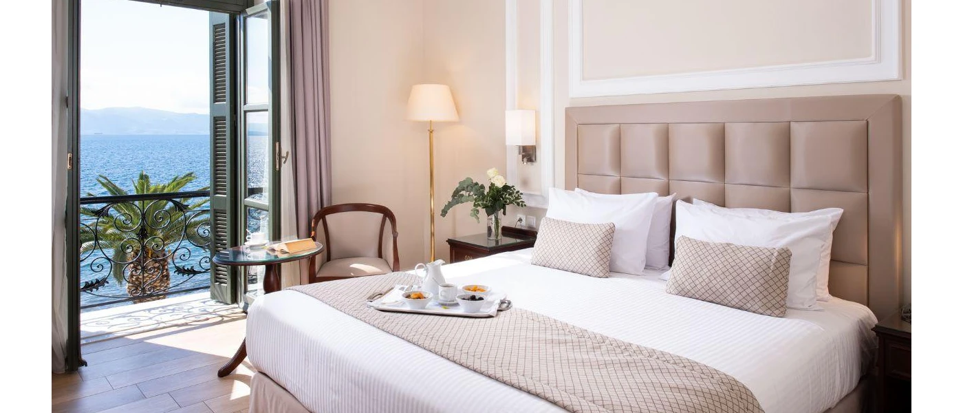 Hotel room with a cream wall, cream padded headboard, white bedding and neutral cushions and balcony with sea view 