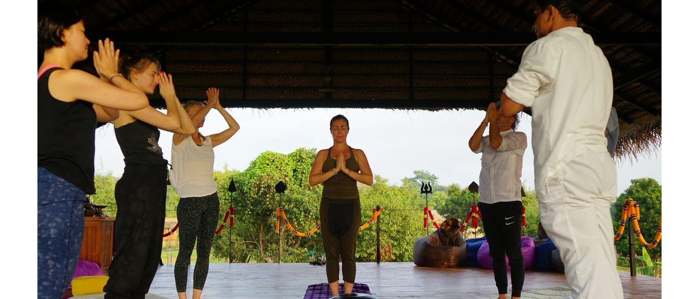 Group in active wear in an open-sided pavilion, with their eyes closed and hands clasped in prayer as an instructor stands at the head of the group