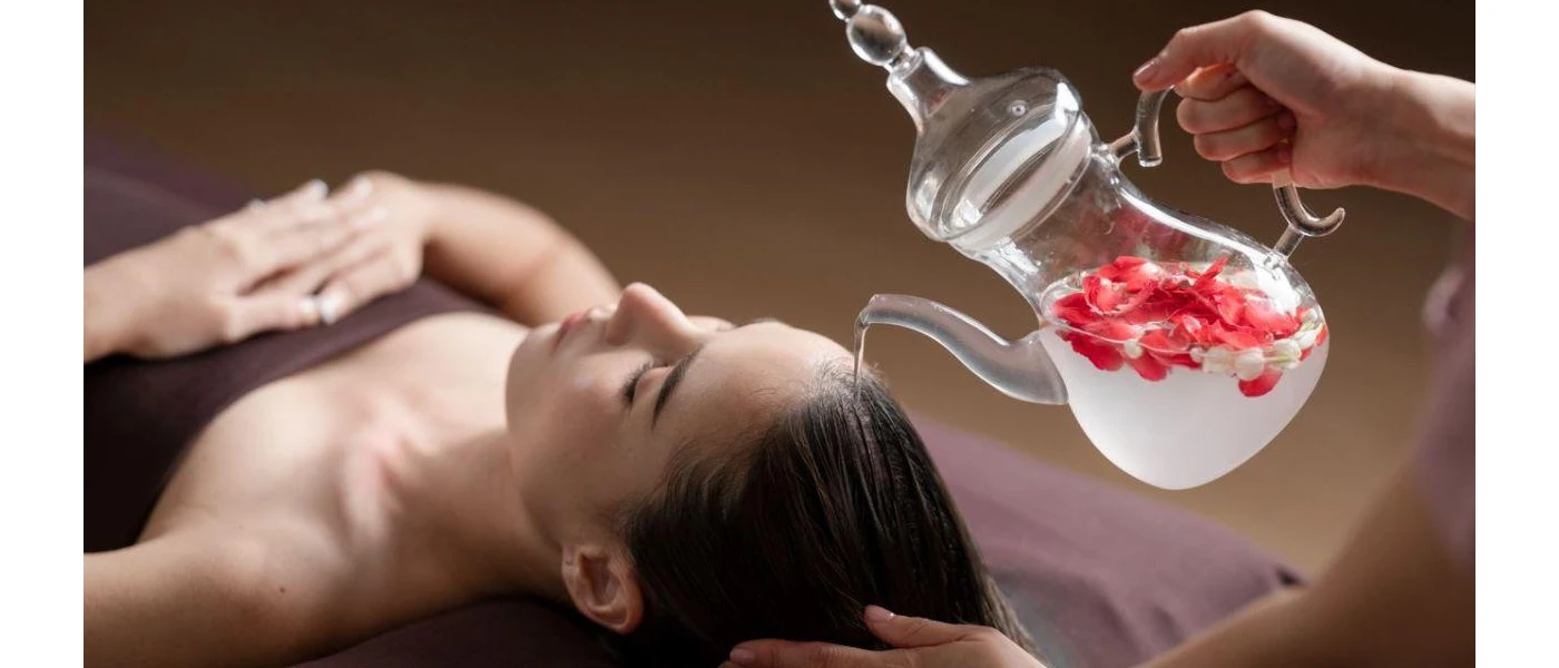 Woman lies on her back with her eyes closed as a therapist pours liquid over her hair from a jug filled with rose petals