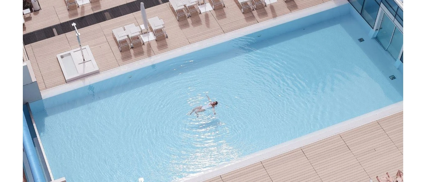 Woman in a white swimsuit floats on her back in a rectangular pool surrounded by wooden decking and loungers