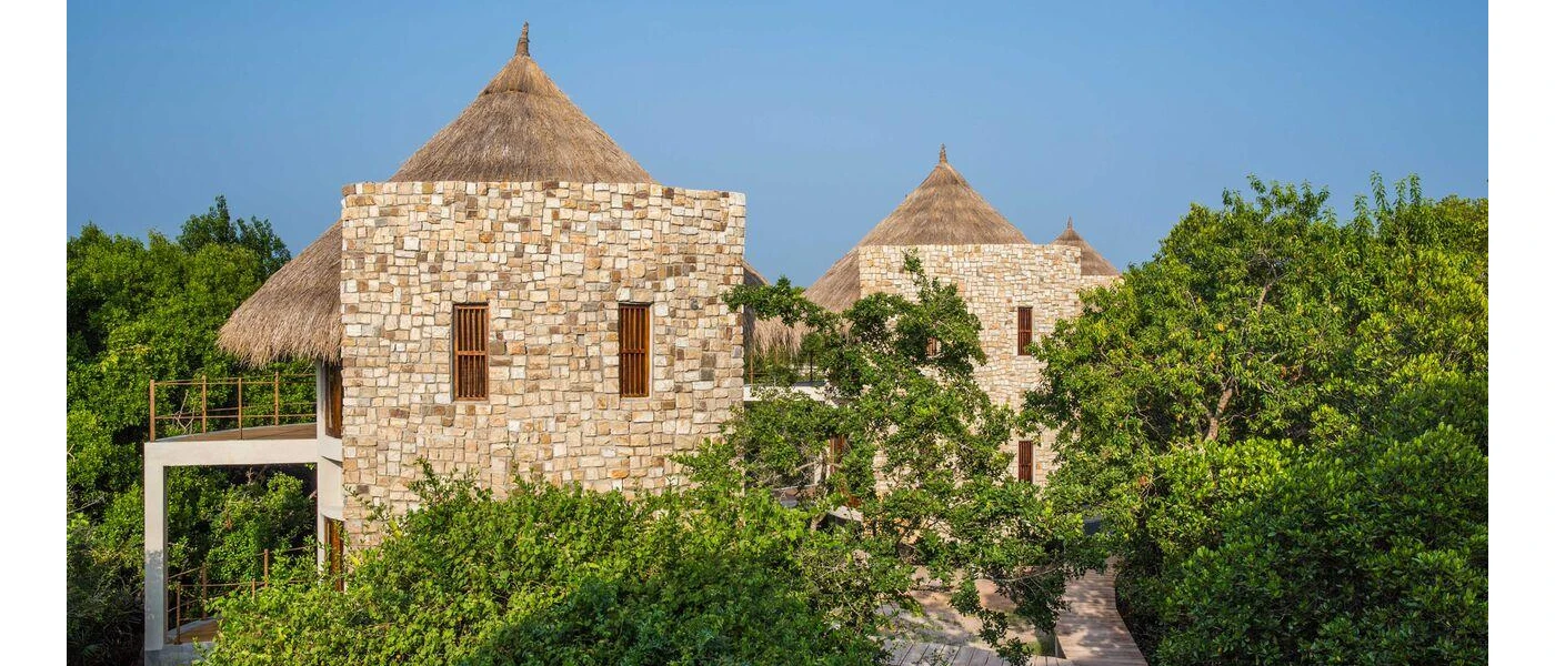 Cobbled stone buildings with triangular thatched rooftops among tropical greenery under a blue sky