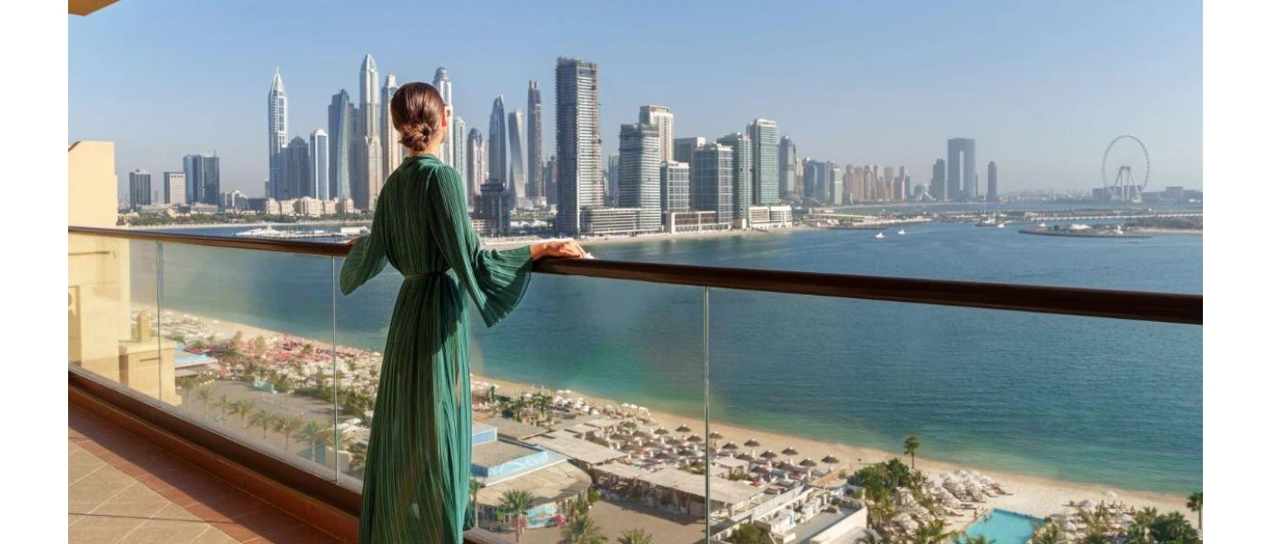 Woman in a green sheer dress looks out over a glass balcony at a white sandy beach, the sea and the Dubai skyline