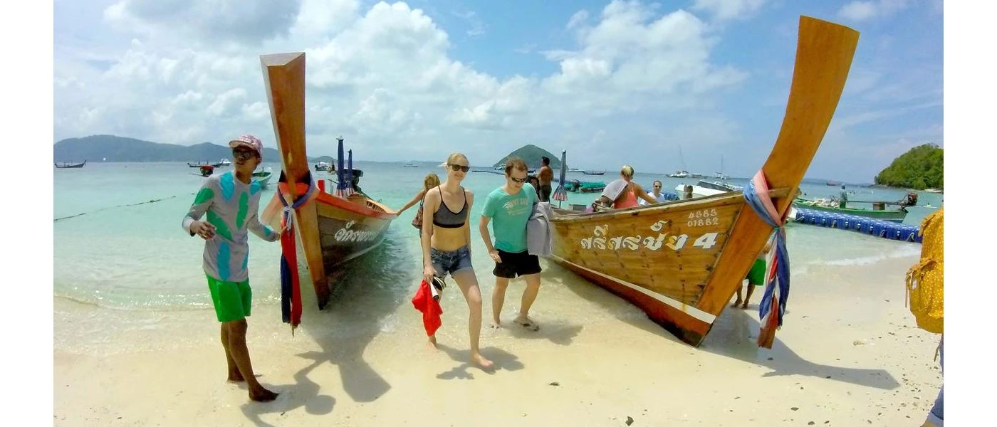 Guests exiting two longtail boats moored on a white sandy beach under a blue sky