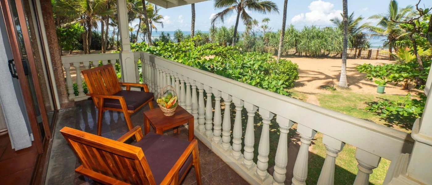 Balcony with wooden chairs and a small table with a fruit basket, with white balustrade overlooking tropical gardens
