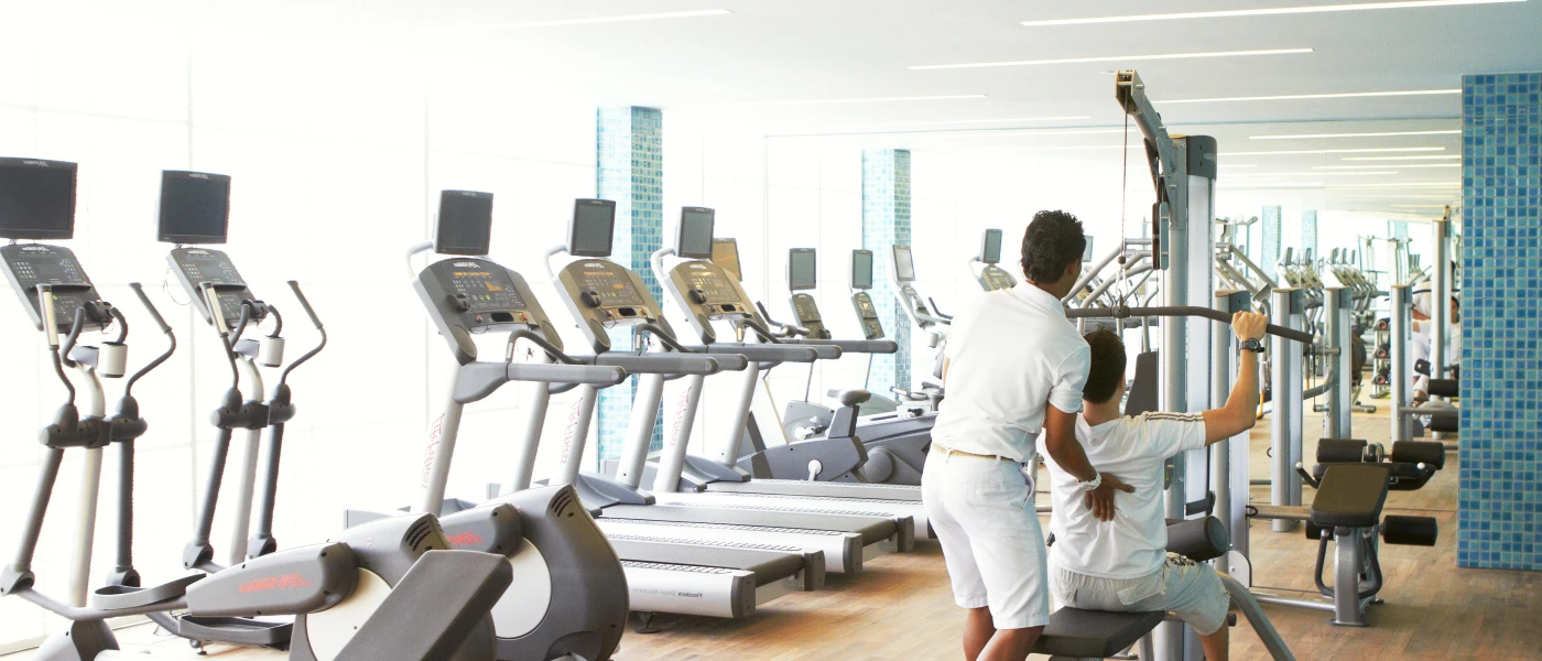 Staff member supports a guest lifting weights in a gym filled with treadmills and step machines