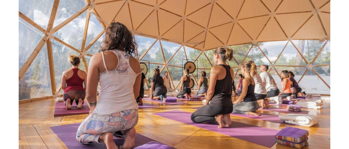 Group in active wear sit on purple mats in a sheer domed building in a class