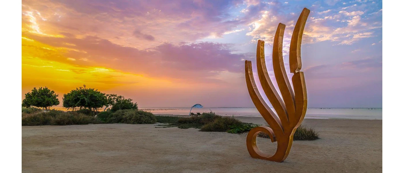 Wave-like sculpture on a sandy beach at sunset, with trees in the background