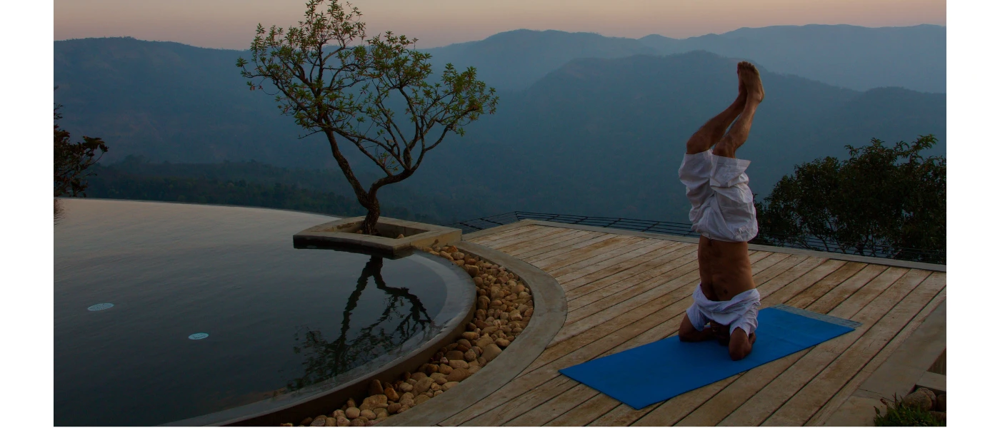 Man in a headstand yoga pose on a blue mat next to an infinity pool with a view of misty hills