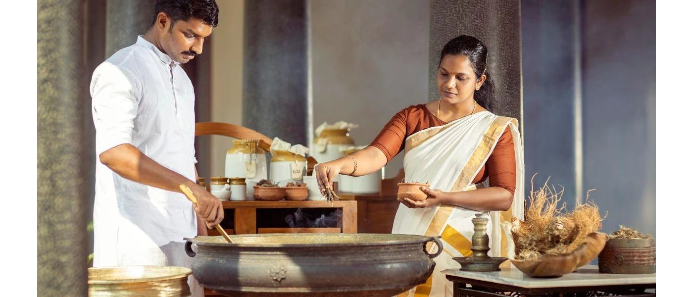 Two staff members in traditional Indian dress work over a large pot surrounded by pots of herbs