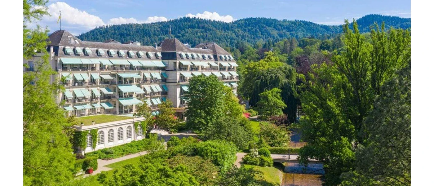 Multi-storey building surrounded by trees, a bridge over water and green-cloaked hills 