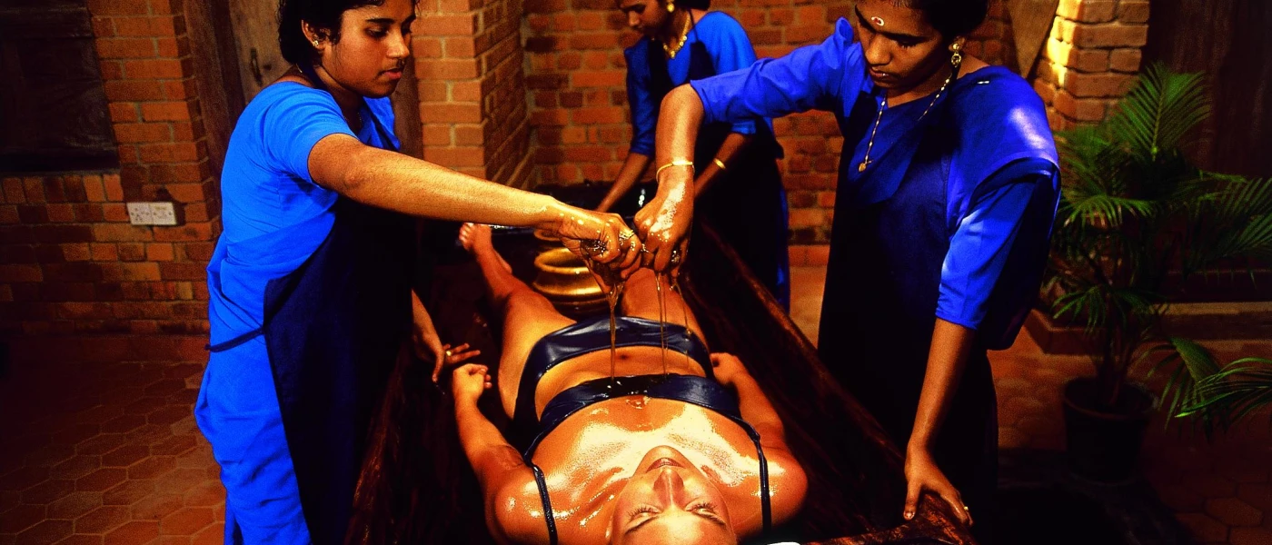 Woman in a black bikini lies on her back on a treatment bed as three therapists in blue uniforms and aprons pour oil over her body