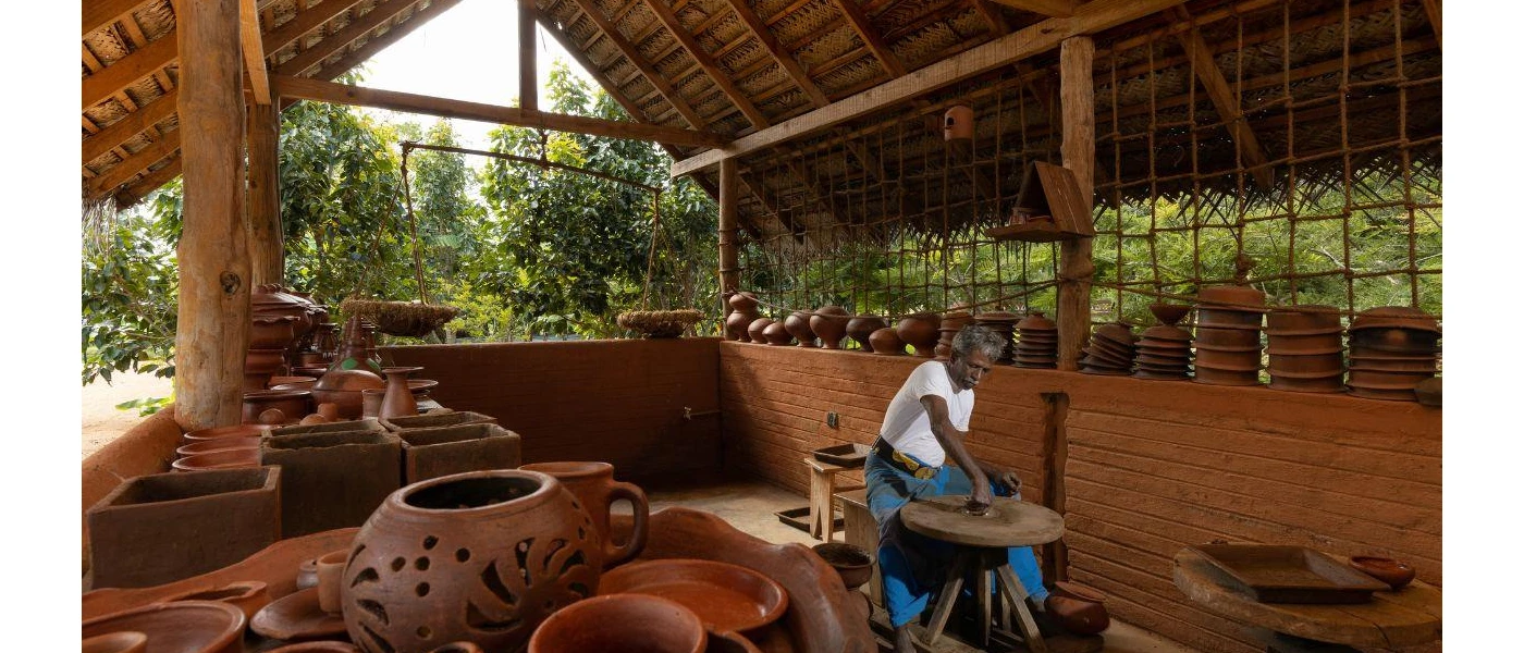 Open-air thatched pavilion filled with terracotta pottery, and a Sri Lankan man shaping a pot on a wooden table in the centre