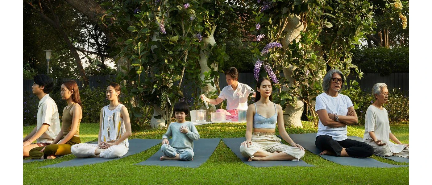 Group of adults and a child sit cross legged with their eyes closed on yoga mats in a tropical garden