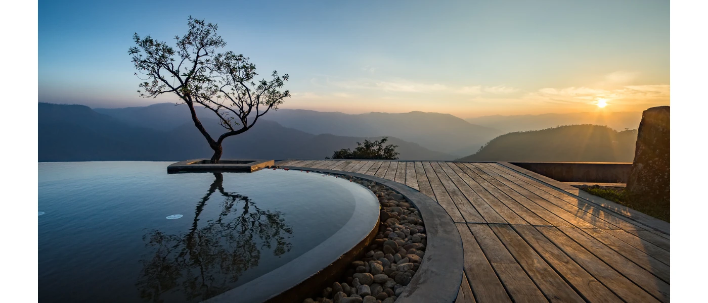 Curved edge of an infinity pool set in wooden decking with a tree next to it and views of misty mountains at sunset