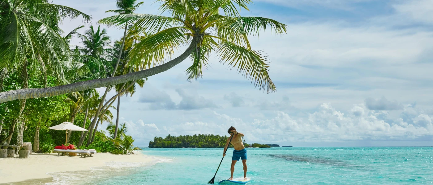 Man in blue swimming trunks stands on a paddle board in turquoise shallows next to a white-sand beach with sloping palm trees