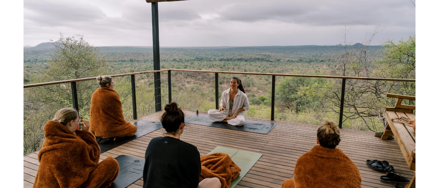 Group on yoga mats in brown blankets sit cross legged on a wooden deck overlooking the African wilderness