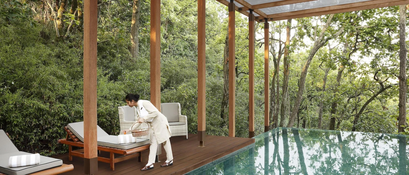 Woman in white traditional dress laying stripy towels on sunbeds next to an infinity pool shrouded by greenery