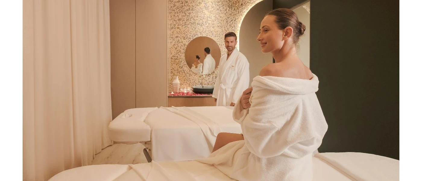 Couple in fluffy white robes in a spa treatment room with white curtains, massage beds with white towels, and a vanity unit covered in pink rose petals