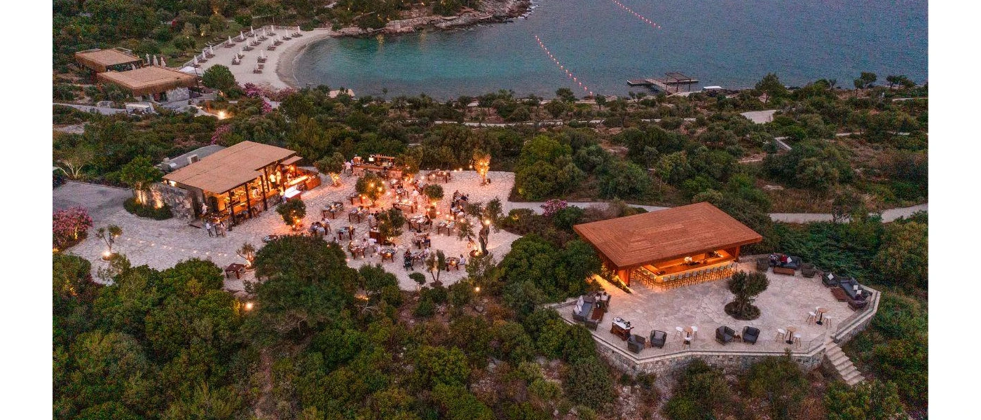 Resort at twilight, with terraces filled with tables and people, surrounding greenery and a neighbouring sandy beach