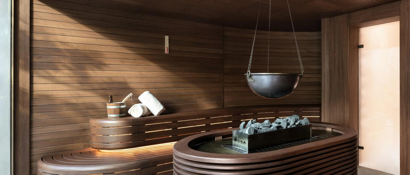 Dark-wood sauna with panelled walls, a wooden bucket and two white towels, and a central table with coal and a bowl tied over the top