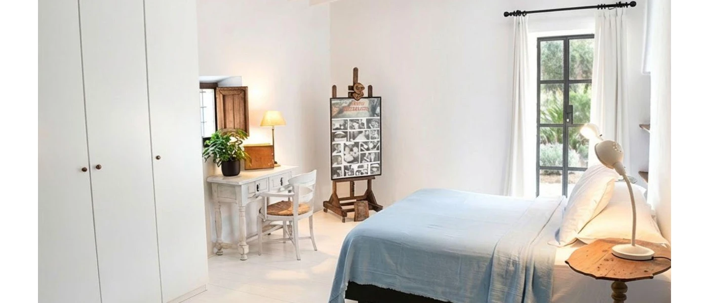 White bedroom with tiled floors, powder blue bedding a white wardrobe and patio doors showcasing gardens beyond