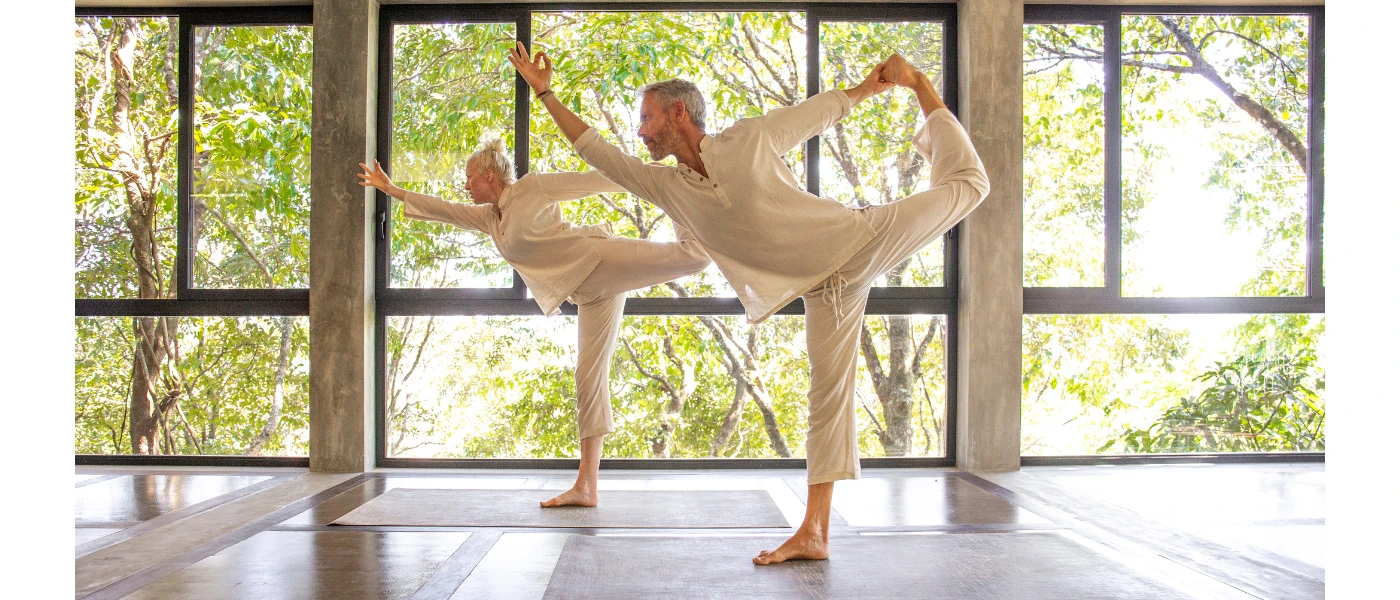 Man and woman in white practice yoga in an airy studio with wraparound windows and tropical greenery beyond