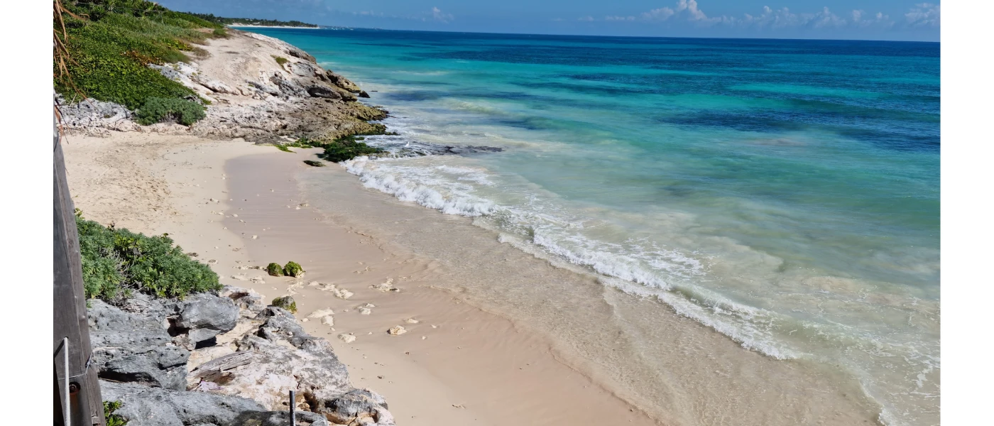 White sandy beach with turquoise shallows backed by rocks and greenery under a blue sky