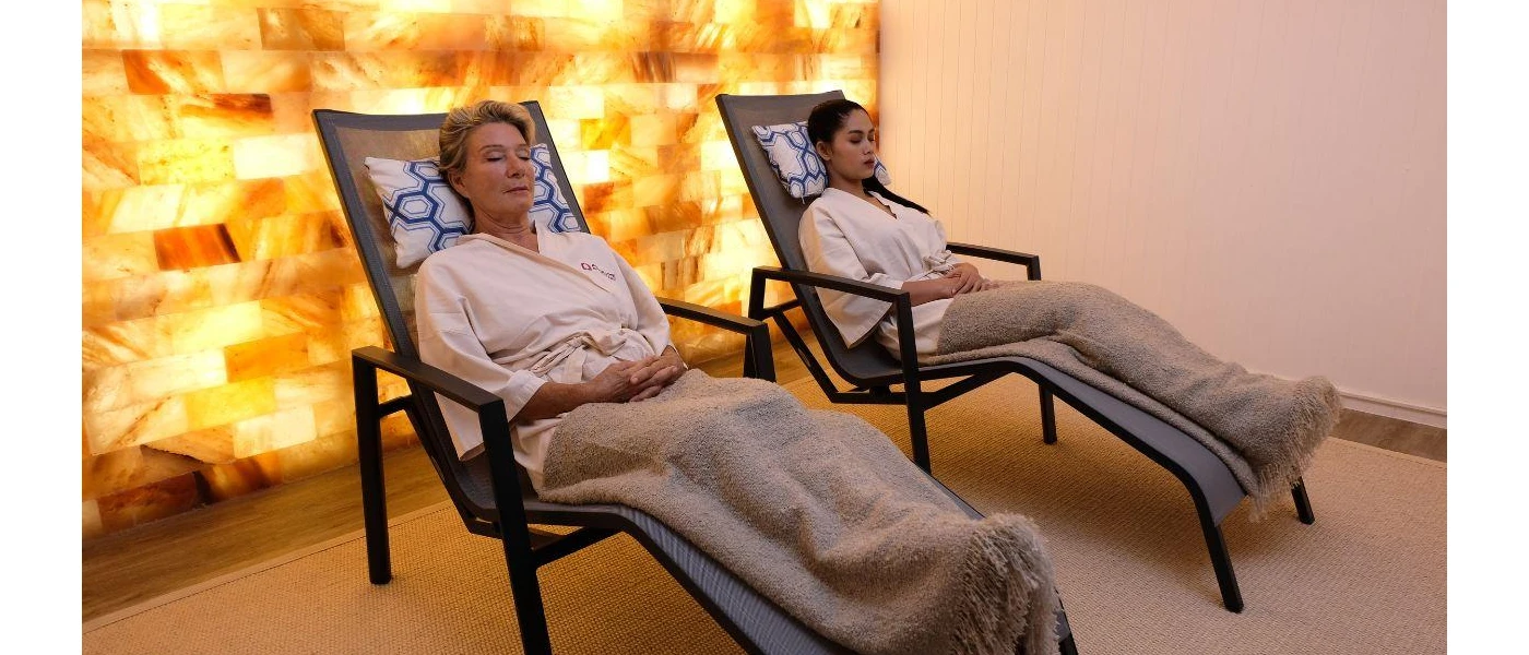 Two women lie on loungers in robes and blankets, with their eyes closed in a room with a gold lit wall