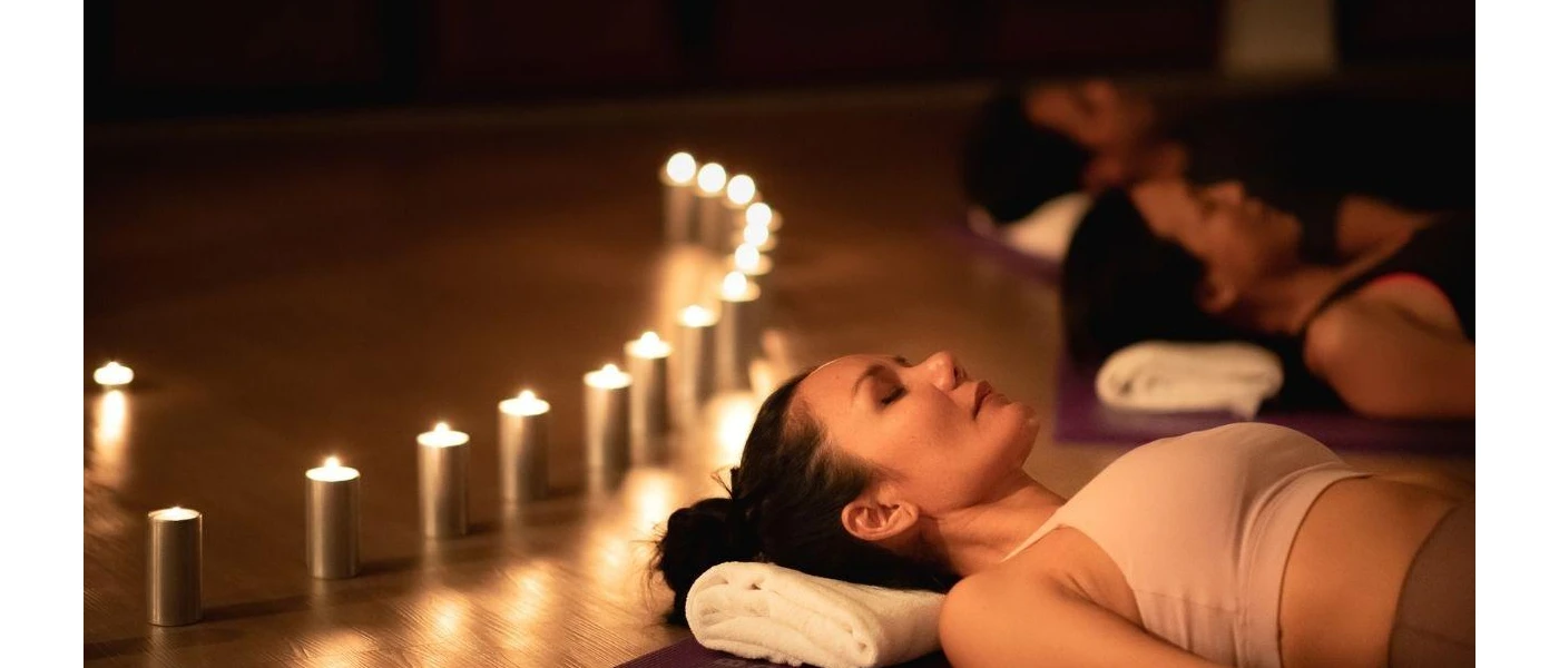 Woman lie on their backs with eyes closed on mats in a dark room lit by candles