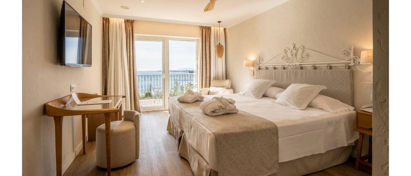 Neutral room with white bedding and cream throw with bathrobes on top, a dressing table and wall-hung TV, and full-length balcony with ocean views