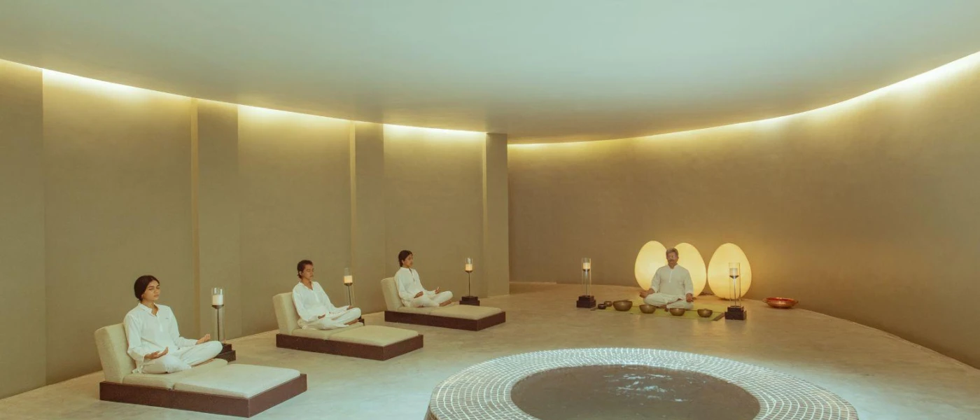 Group of people in white sit cross-legged with their eyes closed on floor cushions in a softly lit cream room as an instructor sits at the front surrounded by gold sound bowls