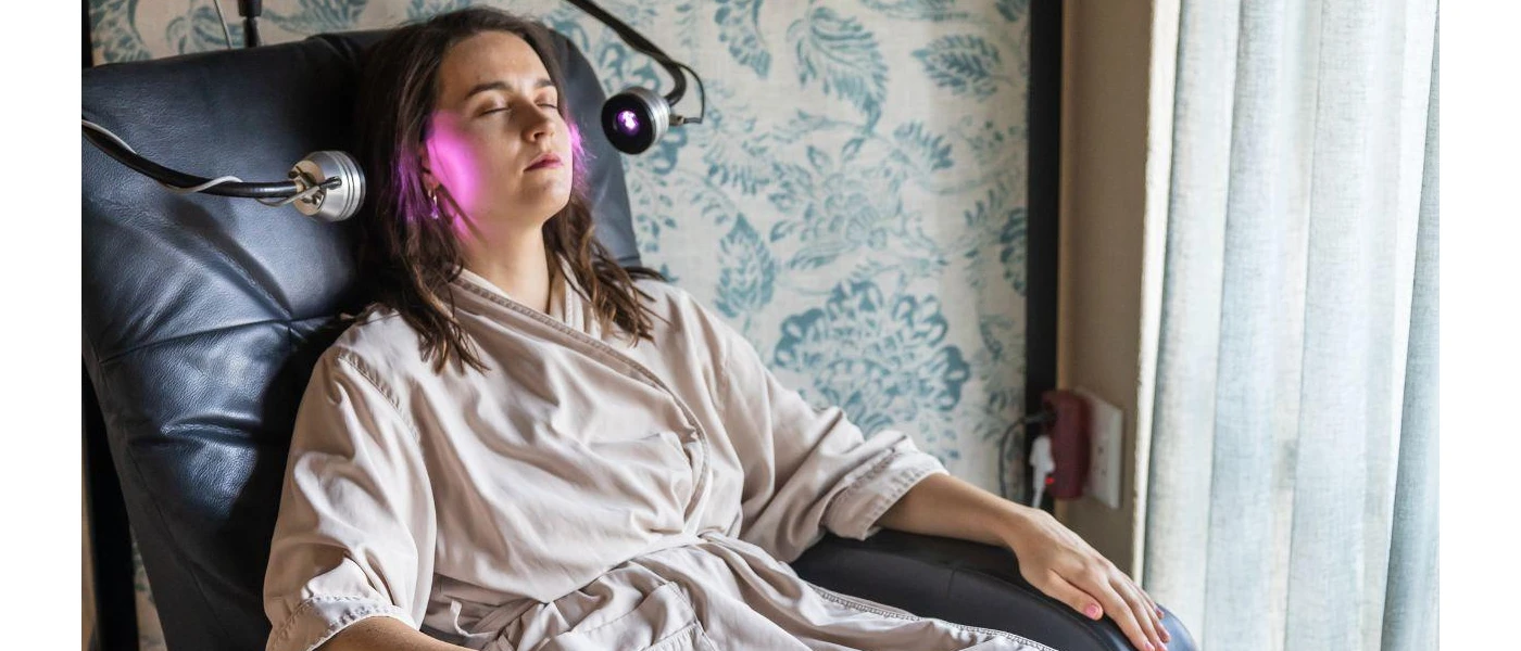 Woman in a beige robe sitting in a black leather chair wearing a head piece, stimulating light therapy