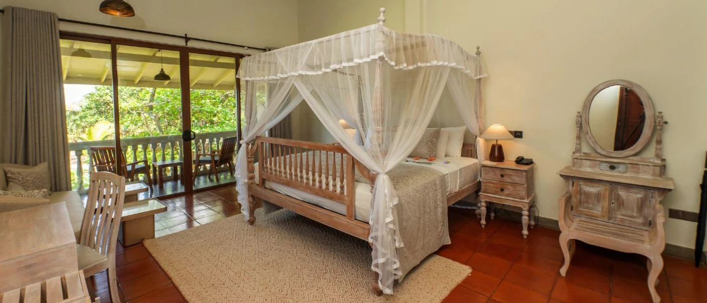 Airy room with a four-poster bed with white drapes, a terracotta tiled floor, desk, dressing table and full-length balcony doors
