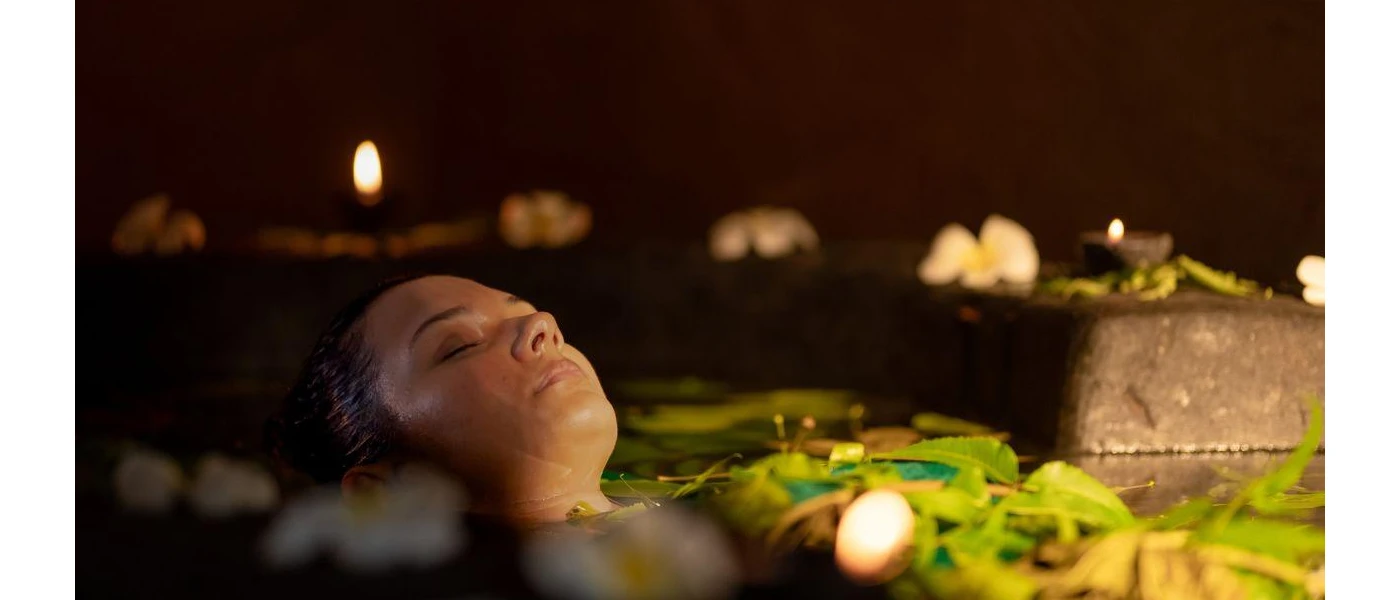 Woman relaxes in a bath filled with plants and flowers in a dark room lit by candles