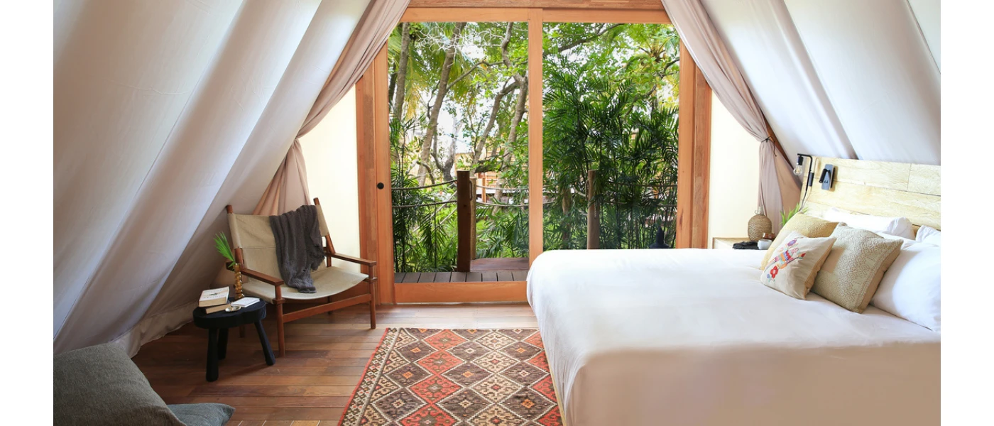 Airy room with inside drapes, white bedding, an aztec-style rug and wooden chair, and full-length balcony doors with a jungle view