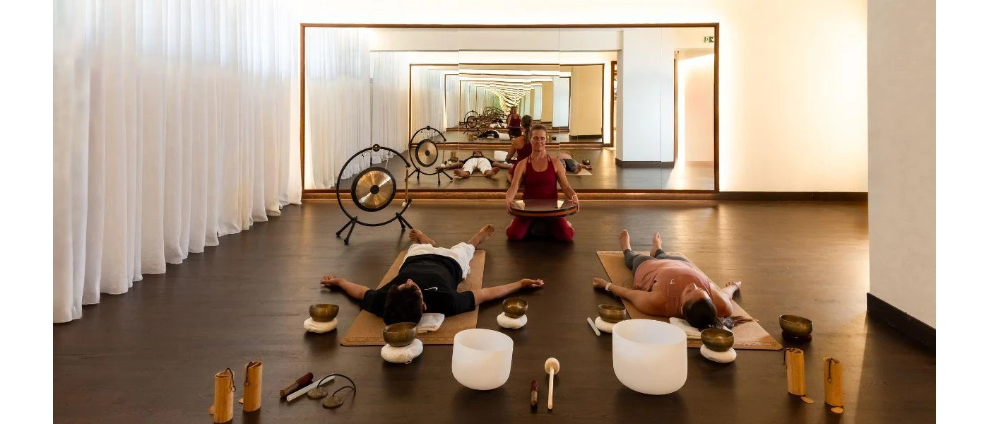 Two people lie on mats surrounded by singing bowls as a woman sitting next to a large gong heads up the class from the front