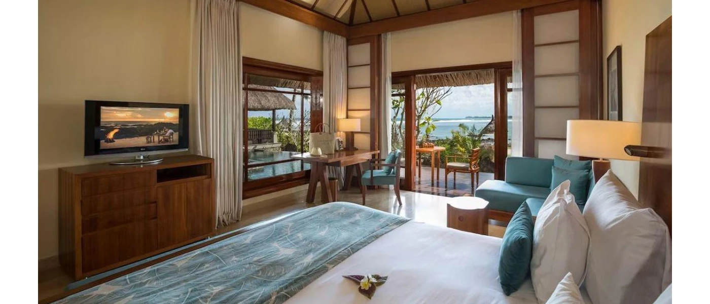 Bohemian-chic hotel room with wooden furnishings and white bedding, with blue cushions, a blue sofa and a blue throw, with a view outside to a furnished terrace and the ocean just beyond