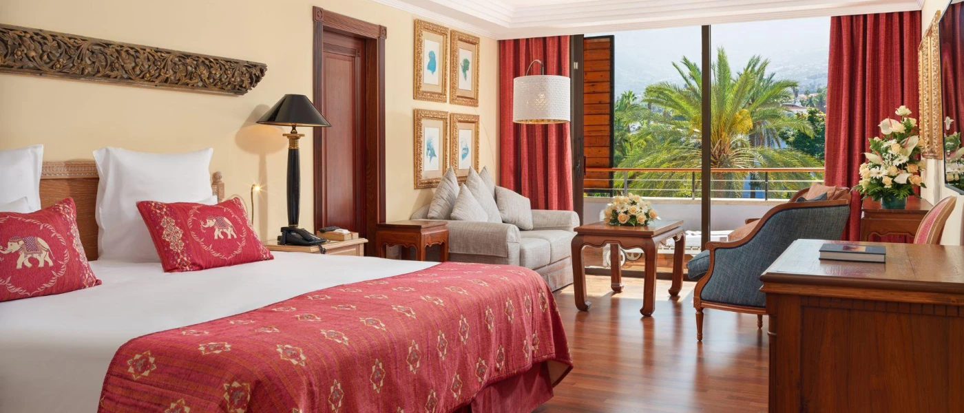 Hotel room with cream walls, white bedding, red soft furnishings, a blue chair and dark-wood furniture