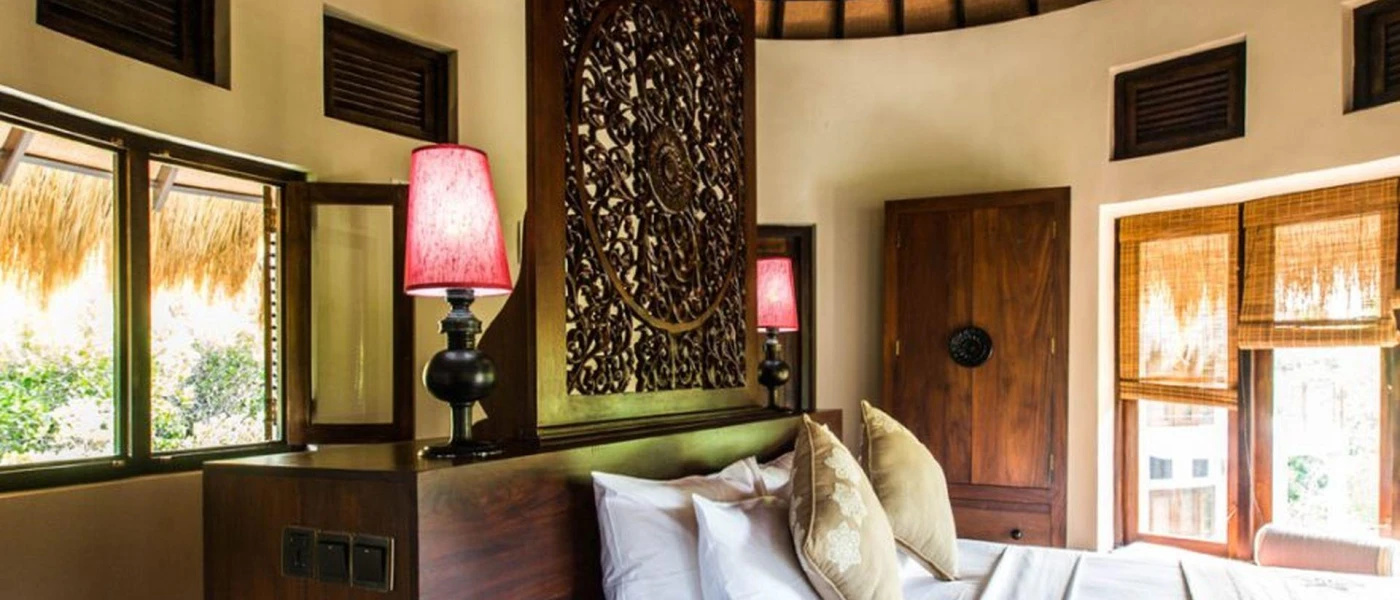 Cabana interior with an intricately carved dark-wood headboard, a wardrobe and white bedding overlooking patio doors with rattan blinds 