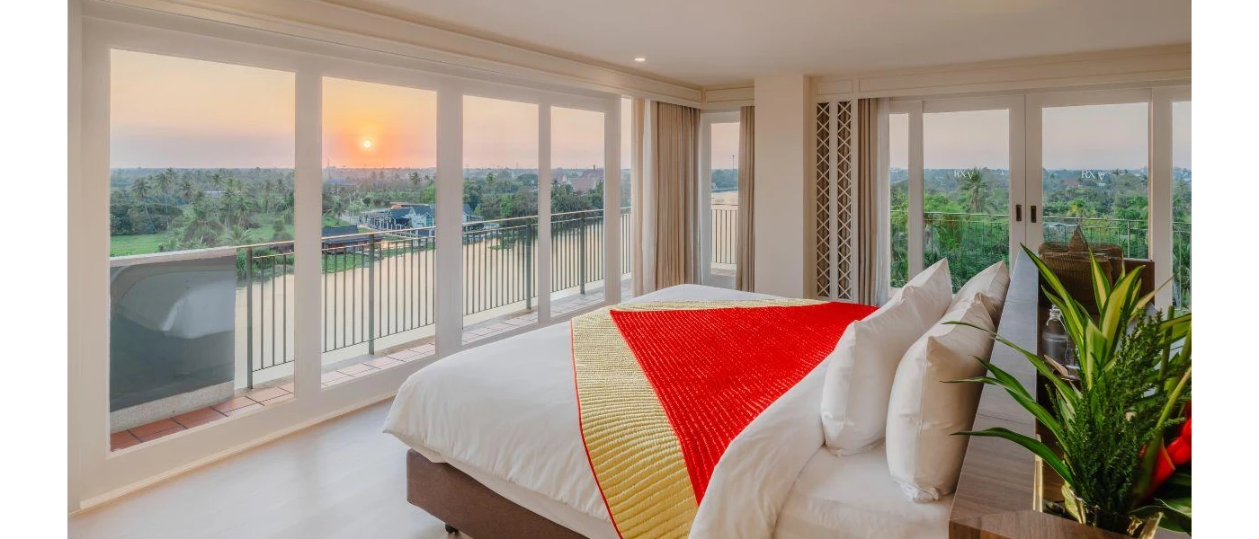 Stylish bedroom with a wraparound balcony with views of surrounding greenery and a lake, with white bedding and a red and gold throw, and a potted green plant in the corner