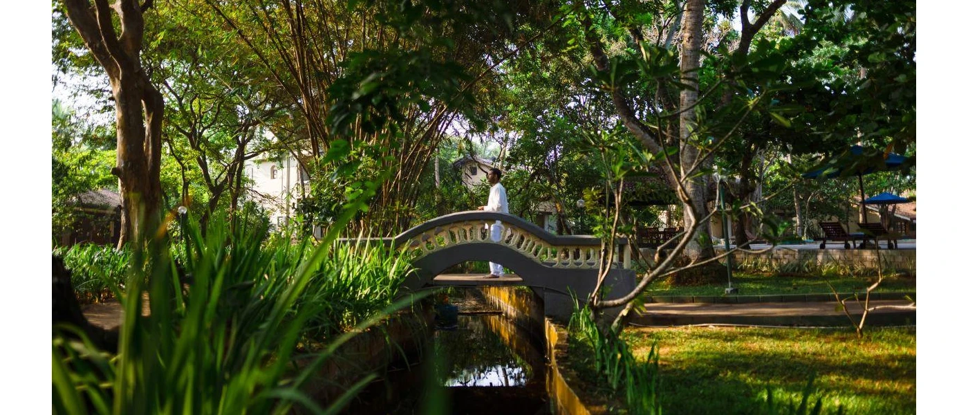 Man in white walks across a bridge over a steam surrounded by tropical gardens