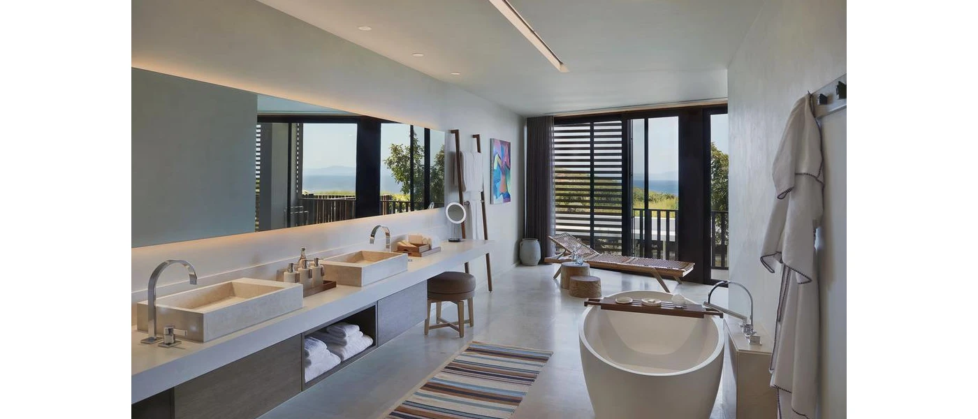 Spacious bathroom with a standalone bath tub, large mirror and twin sinks, a hanging robe and floor-to-ceiling glass with a sea view