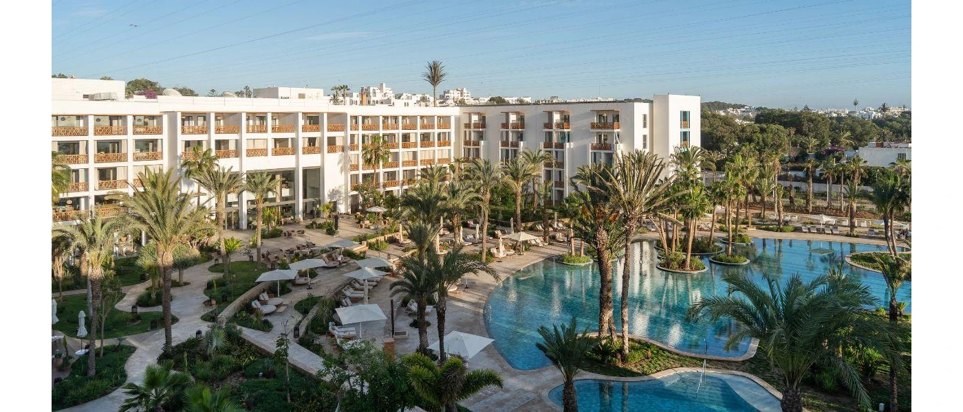 High-rise white hotel overlooking a large swimming pool in grounds covered in palm trees