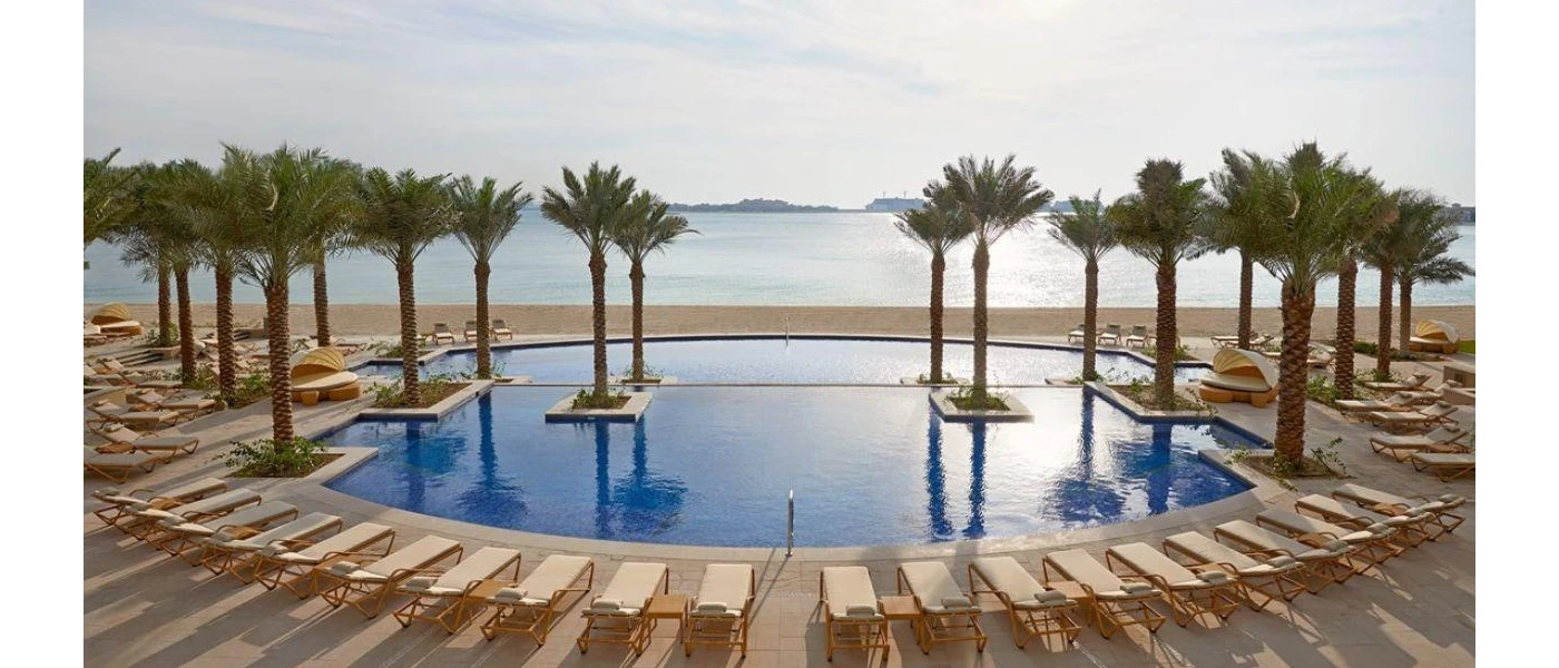 Circular ocean-facing pool lined with white-cushioned wooden loungers and palm trees